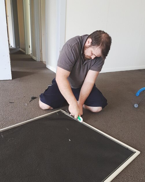 A person installing interlocking rubber flooring tiles in a spare room. The room is partially completed, with some tiles already in place and others ready to be installed. The tiles are black and textured, providing a non-slip surface for workouts.