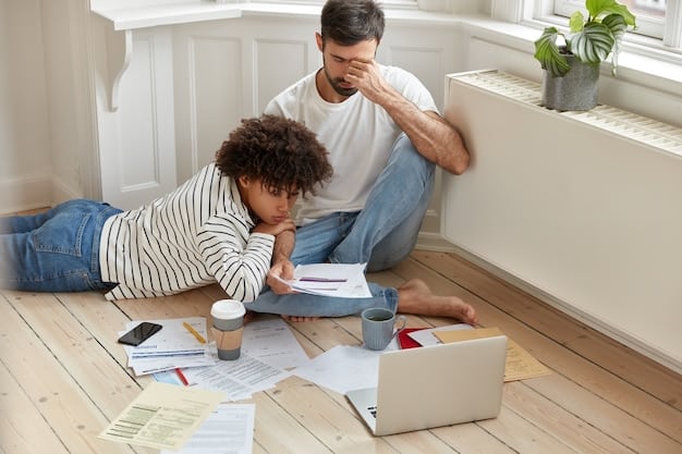 A stressed single father looking at a pile of unpaid bills on his desk at home. His child is sitting at another table.