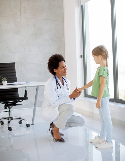 A pediatrician in a white coat smiling while talking with a child and their two parents in an examination room.