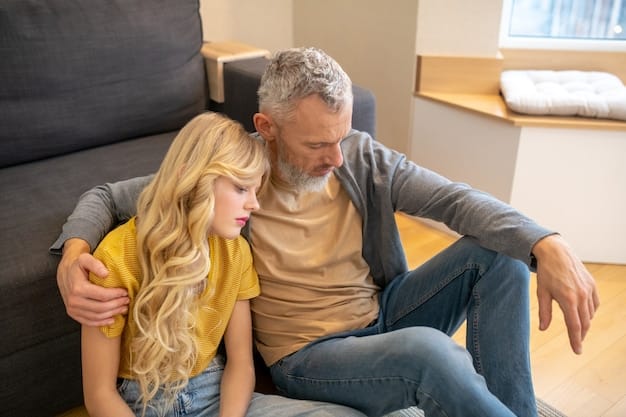 A father comforting his teenage daughter, who is visibly upset. They are sitting on a couch, and the father is holding her hand, showing empathy and support.